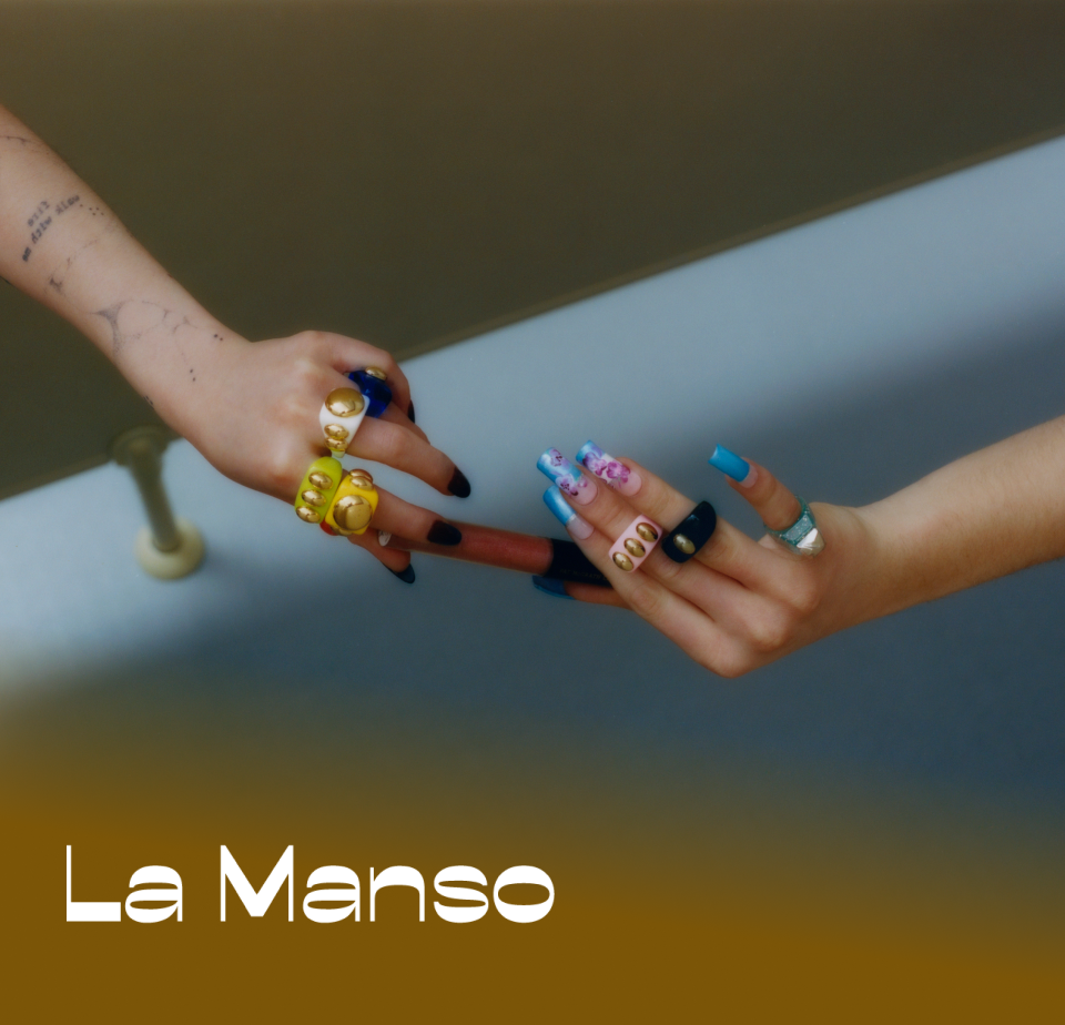 Dos manos se acercan entre sí, adornadas con arte de uñas colorido y anillos llamativos. El fondo presenta colores suaves y difuminados. El texto dice "La Manso."